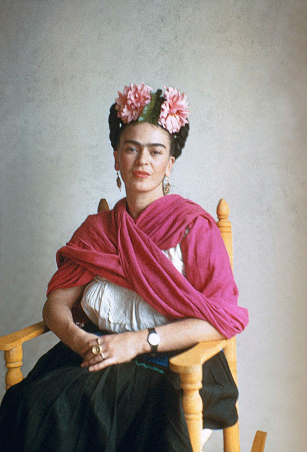 Frida Kahlo seated in photograph with pink flowers in her hair and a bright pink shawl